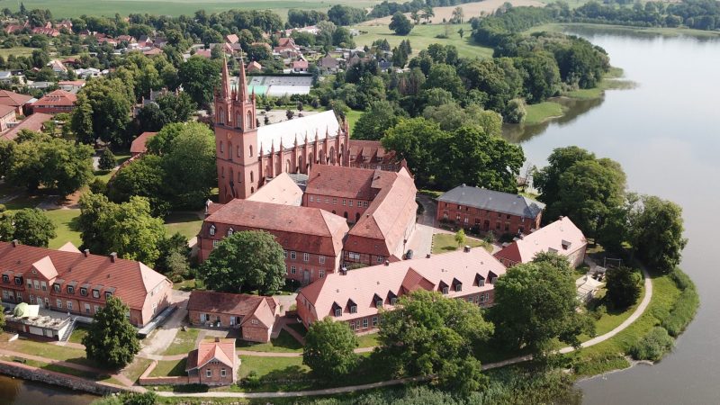 Luftaufnahme der Klosteranlage mit der zweitürmigen Klosterkirche in Dobbertin und weiteren Häusern umgeben von Feldern und Wäldern und dem Dobbertiner See