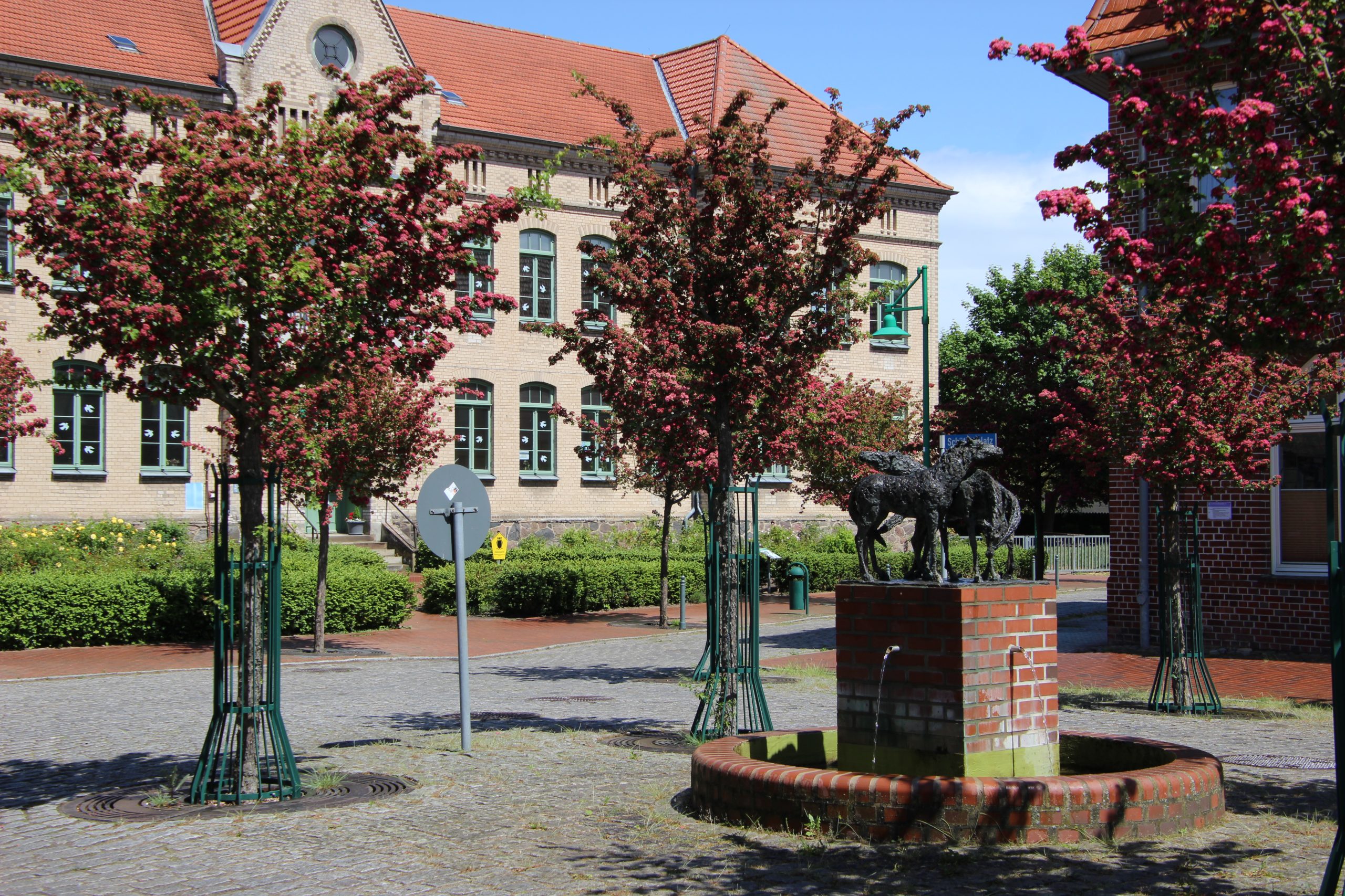 zweigeschossiges Gebäude der Grundschule John-Brinkman in Goldberg davor Bäume und eine Brunnenskulptur mit drei Pferden