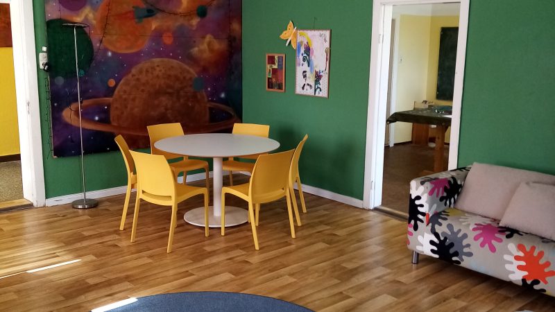 Raum mit runden Tisch und gelben Stühlen, zwei bunte Sofas, ein runder blauer Teppich und bunten Bildern an den Wänden
